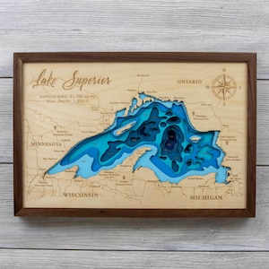 May include: A framed wooden bathymetric map of Lake Superior featuring layered blue cutouts representing water depth. The light wood surface displays engraved text including Lake Superior, surface area, max depth, and regional locations.