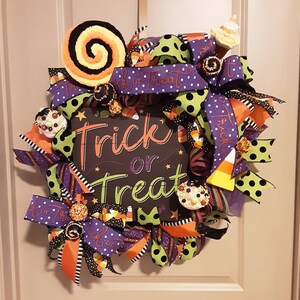 May include: A Halloween wreath with a black and white sign that says "Trick or Treat". The wreath is decorated with purple, green, orange, and black ribbons, candy corn, and other Halloween-themed decorations.