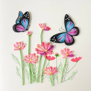 May include: Paper quilling art featuring two butterflies with blue and pink wings, surrounded by pink and white cosmos flowers with green stems.
