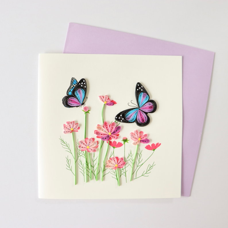 May include: A handmade greeting card featuring a 3D paper craft design of pink and purple cosmos flowers with two butterflies, one blue and one purple, against a white background.