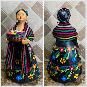 May include: A colourful ceramic figurine of a woman wearing a black dress with floral designs, a striped shawl, and a crown. She is holding a basket of fruit.