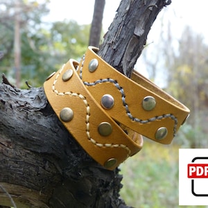 Op de afbeelding: Twee mosterdgele leren armbanden met decoratieve stiksels en zilveren studs. De armbanden zijn om een ​​tak gewikkeld.