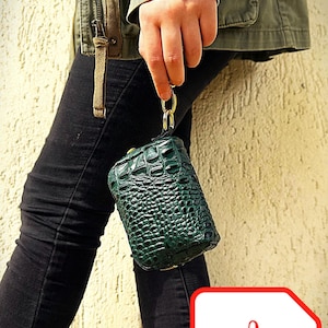 Peut inclure: Une pochette rectangulaire vert foncé texturée avec un fermoir doré et une dragonne assortie. La pochette est tenue par une personne portant une veste verte et un pantalon noir. Une icône PDF est dans le coin inférieur droit.