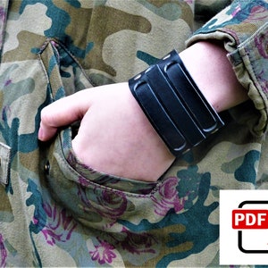 Peut inclure: Bracelet manchette en cuir noir avec trois bandes parallèles et détails de rivets. Le bracelet est porté sur un poignet, avec une veste de camouflage en arrière-plan. La veste a un motif floral.