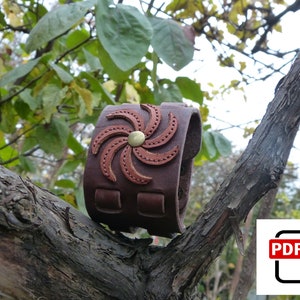 Puede incluir: Un brazalete de cuero marrón con un diseño de estrella roja en cuero y un botón de metal dorado. El brazalete está en una rama de árbol.