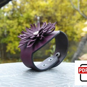 Puede incluir: Un brazalete de cuero morado con un gran diseño de flor detallado. El brazalete es ajustable y tiene un cierre de presión.