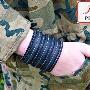 Peut inclure: Bracelet manchette en cuir noir avec coutures blanches. Le large bracelet est composé de plusieurs bandes et se porte au poignet. Le fond est une veste de camouflage.