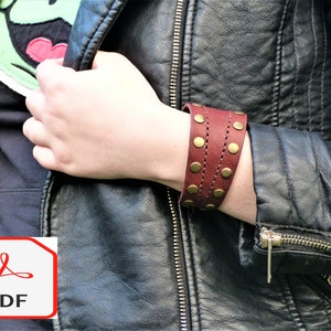 May include: A wide, brown leather cuff bracelet with gold-toned metal studs and stitching. The bracelet is worn on the wrist of a person wearing a black leather jacket.