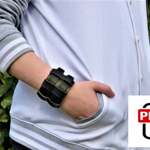 Puede incluir: Un brazalete de cuero negro con un diseño texturizado. El brazalete se lleva en la muñeca.