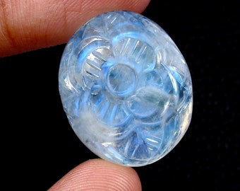 Puede incluir: Un cabujón ovalado de piedra lunar tallada con un diseño floral. La piedra es de color azul claro con destellos iridiscentes.
