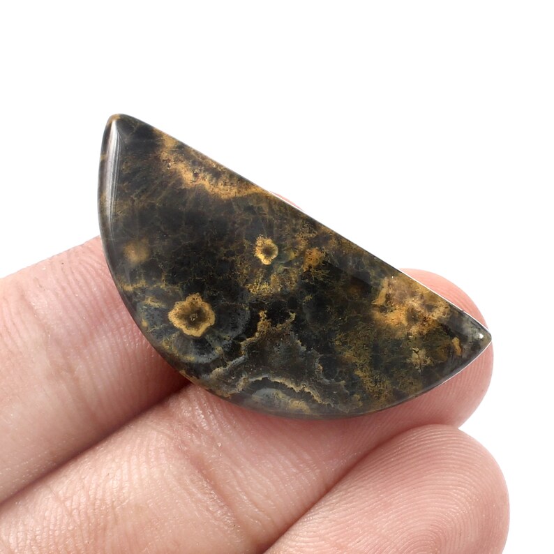 May include: A half-round cabochon of black and gold dendritic agate. The agate has a swirling pattern of black and gold dendrites.