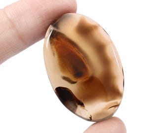 May include: An oval-shaped gemstone with a polished surface. The stone features a blend of tan, brown, and black hues, with intricate patterns and veining. The gemstone is held between two fingers, showcasing its size and shape.