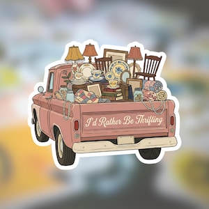 May include: A pink vintage pickup truck sticker filled with thrifted treasures. The truck bed is overflowing with lamps, chairs, framed pictures, teacups, plates, and blankets. The text "I'd Rather Be Thrifting" is on the truck.