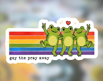 Könnte beinhalten: Ein rechteckiger Aufkleber mit drei grünen Fröschen mit hellgrünen Bäuchen vor einem horizontalen Regenbogenstreifen. Die Frösche lächeln, zwei zeigen das Friedenszeichen. Darüber ist ein kleines rotes Herz und der Text "gay the pray away".