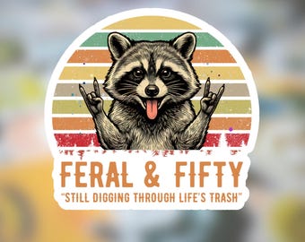 May include: A sticker featuring a raccoon with a rock-and-roll hand gesture, set against a retro sunset design. The text "FERAL & FIFTY" and "STILL DIGGING THROUGH LIFE'S TRASH" are displayed in a bold font.