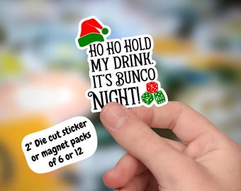 May include: A red and green Christmas-themed sticker with the text "HO HO HOLD MY DRINK. IT'S BUNCO NIGHT!" and a picture of dice. The sticker is 2 inches in diameter and is available in packs of 6 or 12.