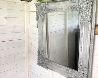 Puede incluir: Un espejo grande, adornado, pintado en plata con un marco decorativo cuelga de una pared de madera blanca. El espejo refleja el área circundante.