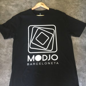Peut inclure: T-shirt noir avec un motif graphique blanc représentant un carré avec des cercles concentriques et le texte "MODJO BARCELONETA".