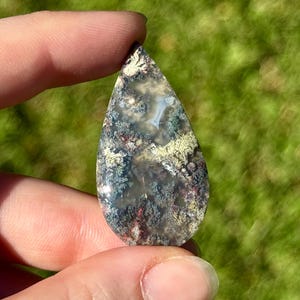 May include: A teardrop-shaped gemstone with a unique pattern of green, red, and white inclusions. The polished surface reveals a landscape-like design, held between fingers against a blurred green background. The stone is approximately 5 cm in length.