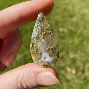 May include: A teardrop-shaped gemstone with a pattern of white, tan, and mossy green inclusions. The polished stone is held between fingers, showcasing its natural beauty and intricate details. The gemstone is a natural, earthy product.