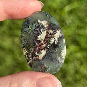 May include: An oval moss agate gemstone. The stone has a translucent green base with detailed patterns of green, black, cream, and reddish-brown inclusions, like a miniature landscape. The stone is held between two fingers against a blurred green background.