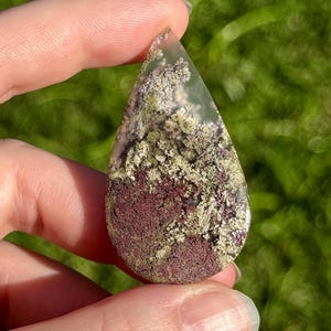 May include: A teardrop-shaped gemstone pendant with a clear, translucent top and a textured interior. The interior features a mix of burgundy, olive green, and cream-coloured patterns. The pendant is held against a blurred green background.