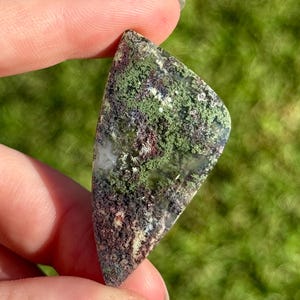 May include: A polished gemstone with a moss agate pattern. The stone has an irregular shape and displays a mix of green, brown, and white hues. The background is a blurred view of green grass.