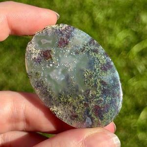 May include: An oval-shaped, polished stone with a mix of colours. The stone features a base of pale green with areas of dark red and patches of mossy green and tan. The stone is held in a person's hand against a blurred green background.