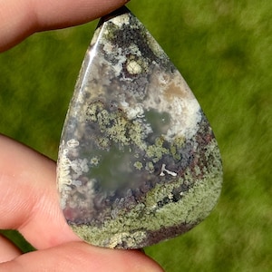 May include: A teardrop-shaped gemstone pendant with a polished surface. The stone features a blend of colours, including green, brown, white, and purple, with a moss-like pattern. The pendant is held between two fingers against a blurred green background.