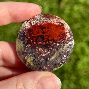 May include: A circular, polished stone displaying a pattern of red, brown, white, and green colours. The stone has a textured surface, with a prominent red central area. The stone is held between fingers against a blurred green background.