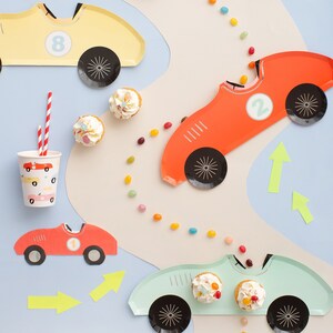 May include: A set of four race car themed paper plates, two red, one yellow, and one blue. The plates feature black wheels and numbers 1, 2, and 8. The plates are surrounded by colourful candy sprinkles and cupcakes.
