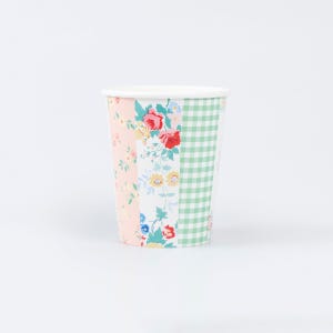 May include: A disposable paper cup featuring a patchwork design of vertical panels. The patterns include a light pink section with small flowers, a central white panel with red and yellow floral motifs, and a green and white checkered section.