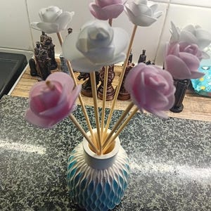 May include: A reed diffuser featuring rose-shaped flowers in white and pink hues. The flowers are arranged in a white and blue patterned vase. The vase sits on a speckled countertop. The flowers are on wooden sticks.