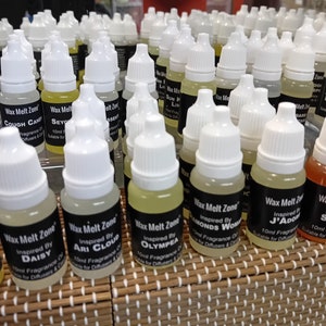 May include: A close-up of a selection of small glass bottles with white caps, filled with a clear liquid. The bottles are labelled with the text "Wax Melt Zone" and various scents, such as "Cough Candy", "Seychelles", "Diamonds Women", "J'Adore", and "Olympia". The bottles are arranged on a brown wicker surface.