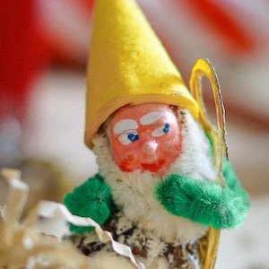 May include: A yellow felt gnome with a white beard and green felt mittens. The gnome is holding a gold shepherd's crook.
