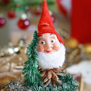 May include: A red and white gnome figurine with a white beard and a pine cone base. The gnome is surrounded by green tinsel and a silver garland.