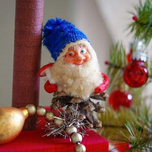 May include: A vintage Christmas ornament featuring a gnome with a blue hat, white beard, and red accents. The gnome is made of various materials and is attached to a pinecone base. A gold and silver beaded garland and a small Christmas tree with red ornaments are also visible.