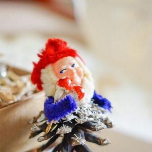 May include: A red and white gnome figurine with a blue hat and a pine cone base. The gnome is holding a red and white mushroom.