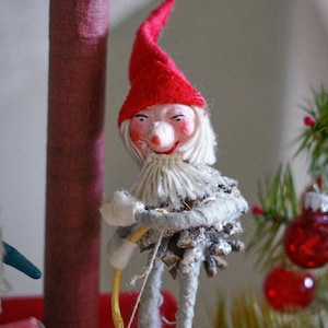 May include: A red and white felt gnome ornament with a white beard and a pine cone body. The gnome is holding a bow and arrow and is wearing red shoes.
