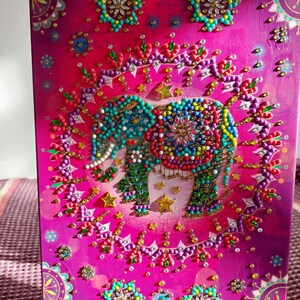 May include: A pink canvas with a beaded elephant design. The elephant is surrounded by a beaded circle with stars and flowers. The canvas is on a wooden easel.