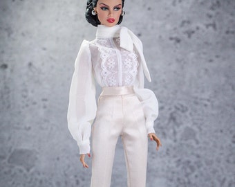 Puede incluir: Una blusa blanca con una superposición de encaje y un lazo en el cuello. La blusa tiene mangas largas y abullonadas. La modelo lleva pantalones blancos y tacones blancos.