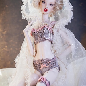 Könnte beinhalten: Eine Modepuppe mit blondem Haar, blauen Augen und rotem Lippenstift, die ein Dessous-Set mit Spitzendetails und rosa Schleifen trägt. Die Puppe trägt auch einen weißen Spitzenschleier.