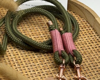 May include: Olive green dog leash with pink accents and rose gold hardware. The leash is made of a thick, braided rope. The leash has a matching pink wrap at the handle and a rose gold clasp. The background includes dried flowers and a woven tray.