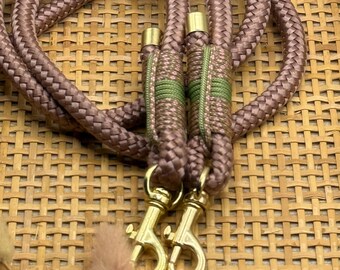 May include: A mauve-colored dog leash with gold-colored metal clasps. The leash is made of a thick, braided rope with green and brown accents near the clasps. The background is a woven, light brown surface.