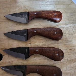 May include: Four handmade knives with dark brown wooden handles and silver rivets. The blades are made of metal and have a slight curve.