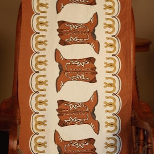 May include: A brown and white table runner with a repeating pattern of brown cowboy boots and gold horseshoes. The runner is on a wooden chair.