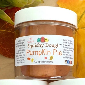 May include: A clear plastic jar with a white lid contains orange-brown Squishy Dough. The label reads "Pumpkin Pie" and "4.5 oz (net weight)". The jar is labeled as non-toxic and contains wheat. The product is a fall special.