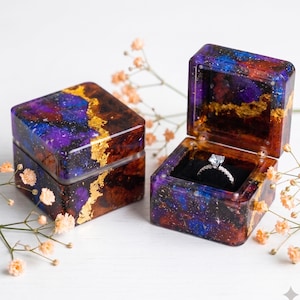 May include: Two square ring boxes made of resin with a galaxy-inspired pattern in shades of deep purple, blue, and black, accented with gold leaf flakes. One box is closed, and the other is open, containing a silver ring with a clear stone.