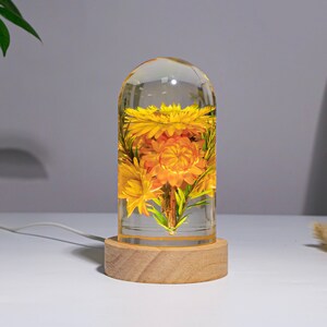 K&ouml;nnte beinhalten: Eine dekorative Lampe mit einem transparenten, kuppelf&ouml;rmigen Glas, das leuchtend gelbe und orangefarbene Blumen umschlie&szlig;t. Die Lampe steht auf einem runden, hellen Holzsockel. Ein wei&szlig;es Kabel erstreckt sich von der Basis.
