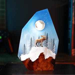 May include: A decorative resin sculpture featuring a moose standing on a snowy mountain under a full moon and starry sky. The base is a dark wood, and the background is a blue and white painted scene. The sculpture is displayed on a black surface.